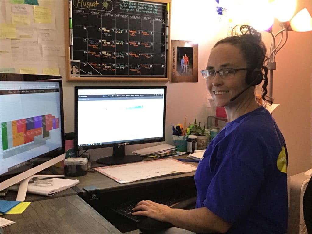 Person wearing a headset sits at a desk with two computer monitors and a large calendar on the wall behind them, smiling at the camera.
