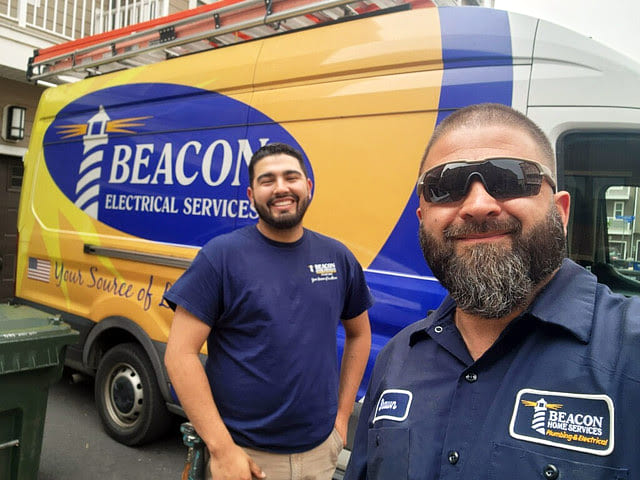 Two bearded technicians pose in front of a Beacon Electrical Services van with a blue and yellow livery, smiling at the camera.