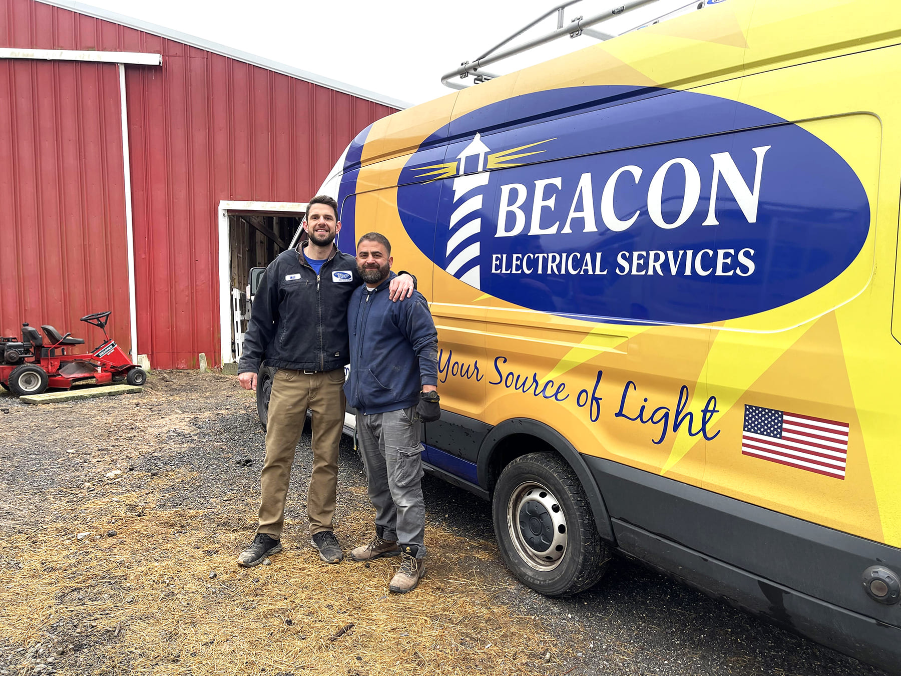 Two electricians pose beside a bright Beacon Electrical Services van with a red barn in the background.