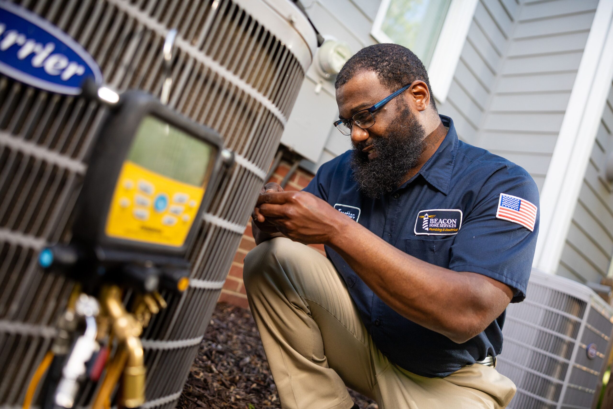 Technician kneeling beside an outdoor air conditioning unit, using tools to repair it in a home setting.