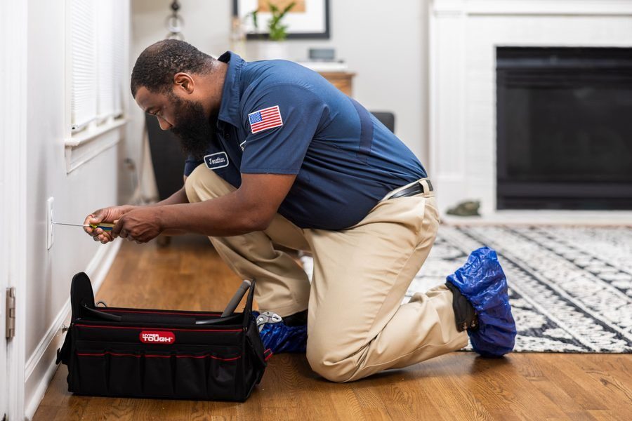 Handyman kneels on a wooden floor, using a screwdriver to work on an electrical outlet with a toolkit nearby in a living room.