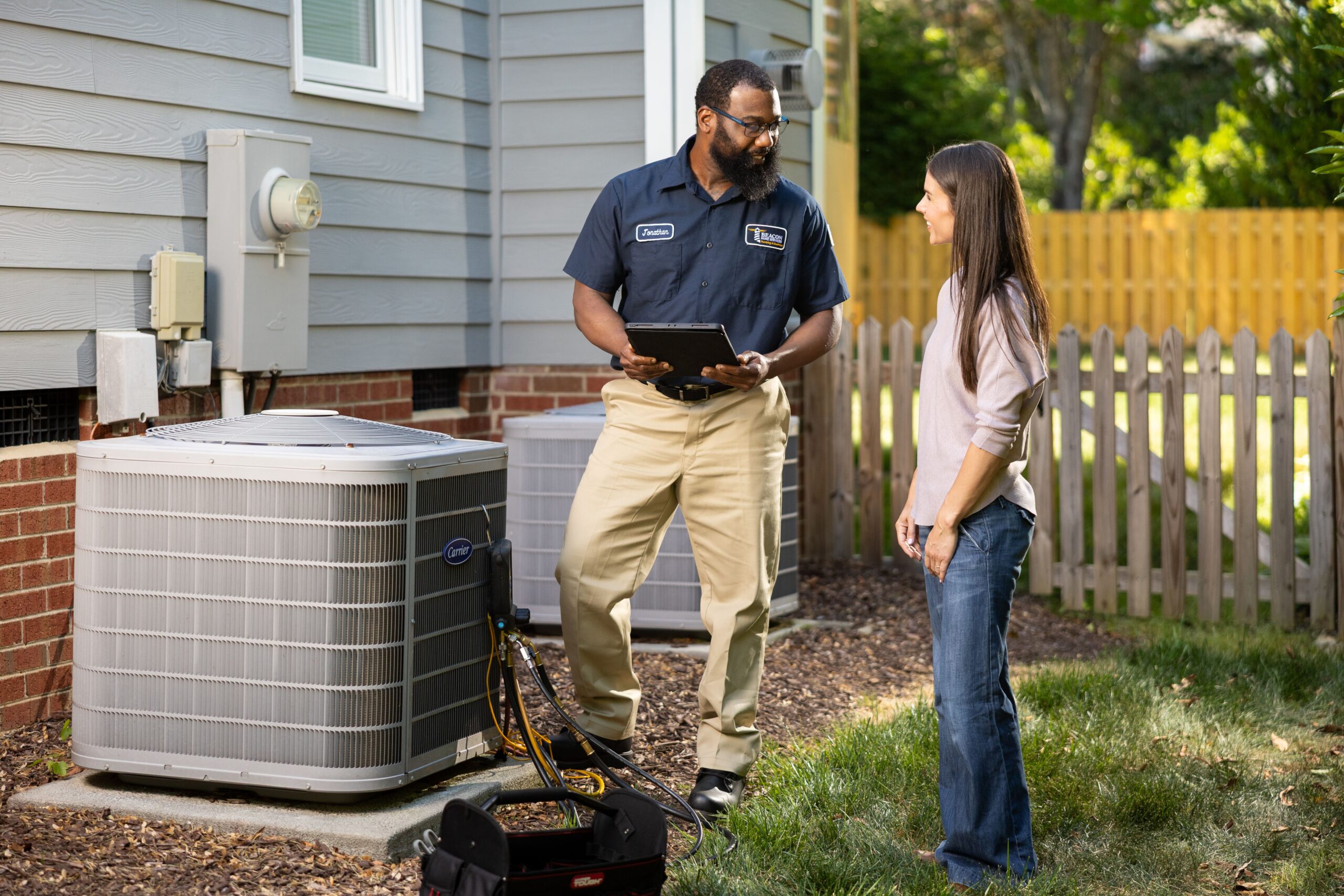 HVAC technician in a navy shirt shows a tablet to a woman beside outdoor air conditioning units outside a house.