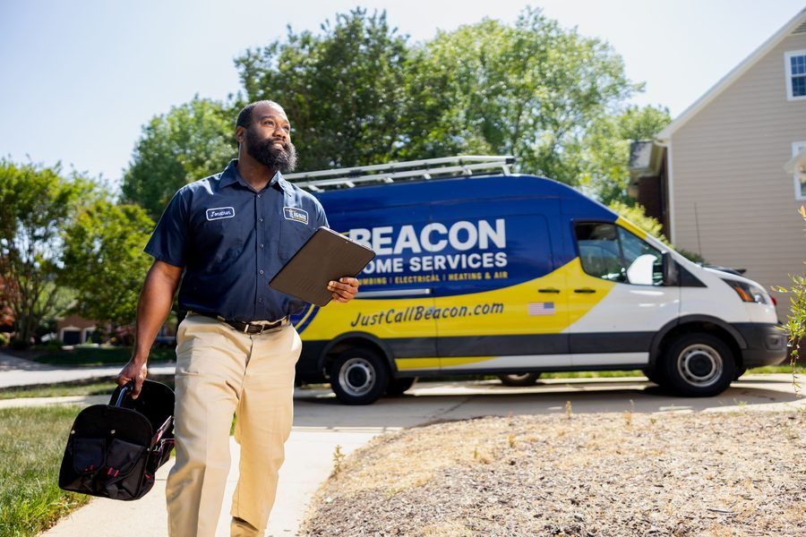 Bearded technician in a navy uniform walks on a sidewalk with clipboard and a tool bag, in front of a blue and yellow Beacon Home Services van.