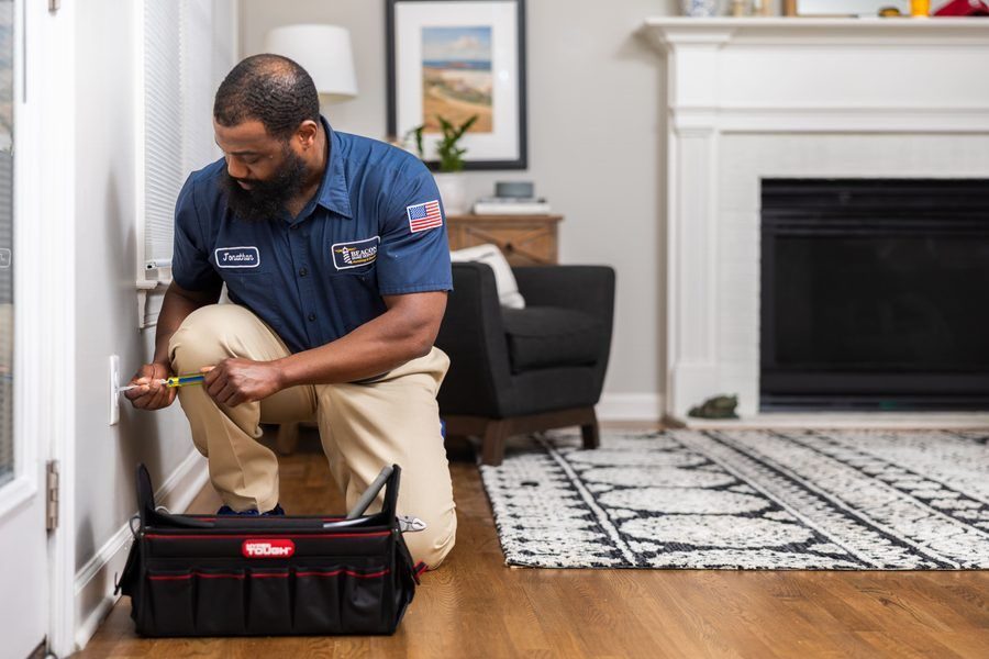 Home repair technician kneels by a wall outlet, screwdriver in hand, with a red toolbox on the floor nearby in a living room.