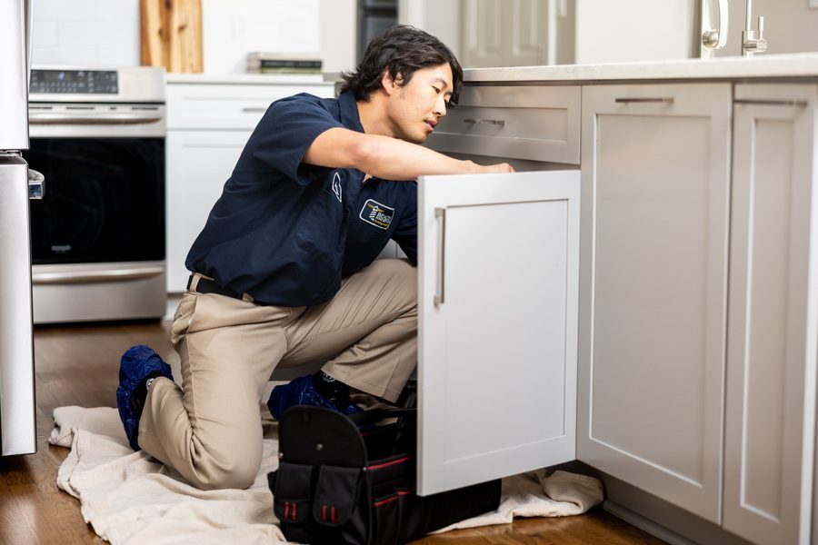 Technician in a navy uniform kneels on a towel, inspecting the inside of a kitchen cabinet under the counter.