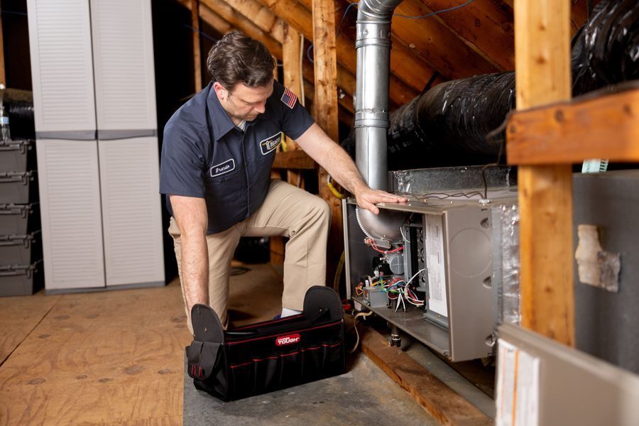 HVAC technician kneels in an attic, inspecting an open furnace with exposed wiring and ducts beside a tool bag.