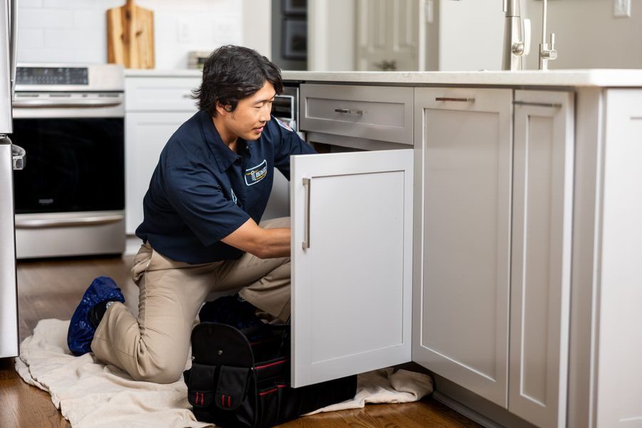 A service technician in a navy polo kneels on a towel under a kitchen sink, opening a lower cabinet to perform repairs with a toolkit nearby.