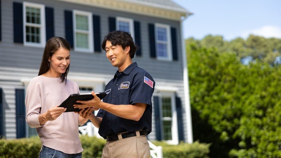 A woman and a man in a navy uniform show a tablet to each other outside a gray suburban home with blue shutters.