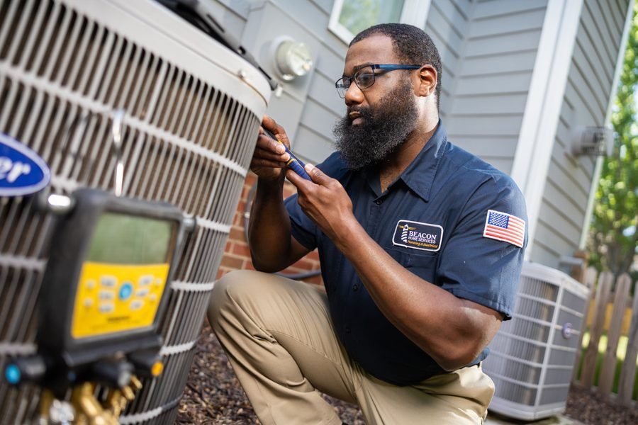 Technician with a beard inspecting an outdoor air conditioner unit using a diagnostic tool, wearing a navy shirt with a patch and American flag patch on the sleeve.