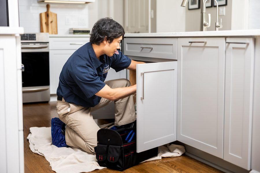 Male technician kneeling on a towel under a kitchen sink, opening a cabinet door with a tool bag beside him.