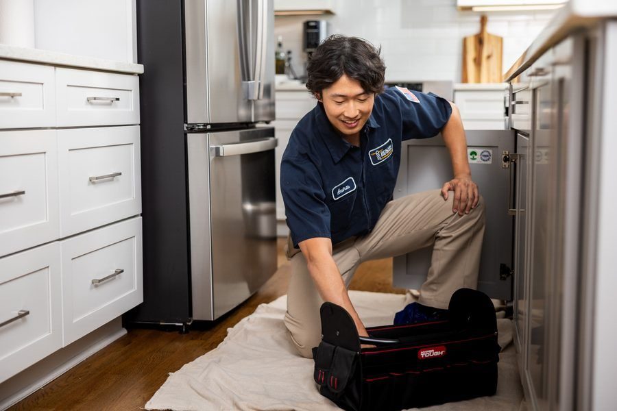 Appliance repair technician kneels on a towel, working under a dishwasher with a tool bag beside him.