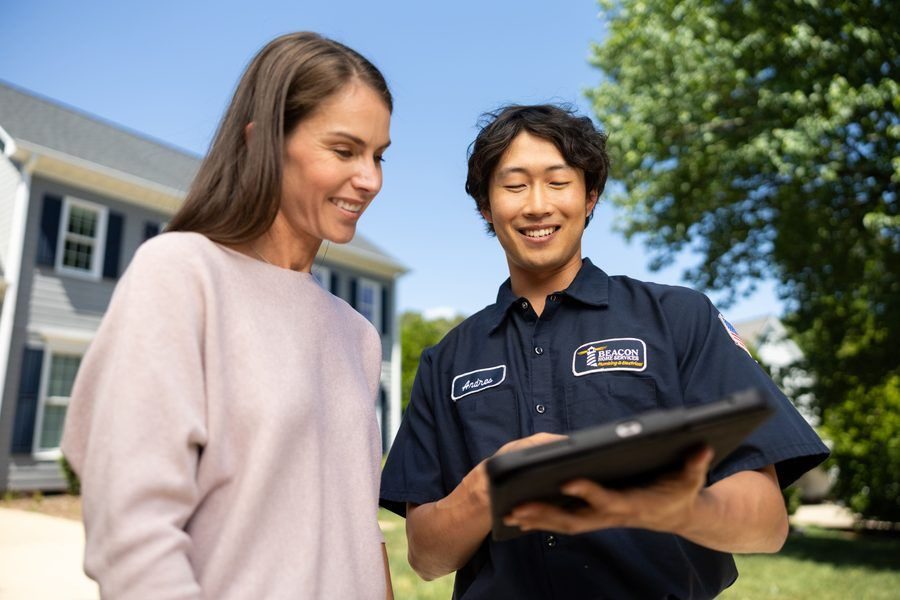 Service technician in a navy uniform shows a tablet to a smiling homeowner outside a suburban house on a sunny day.