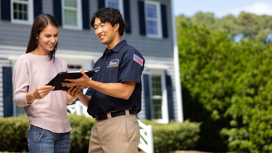 Repair technician shows tablet to smiling woman in front of a suburban house outside a home service visit.