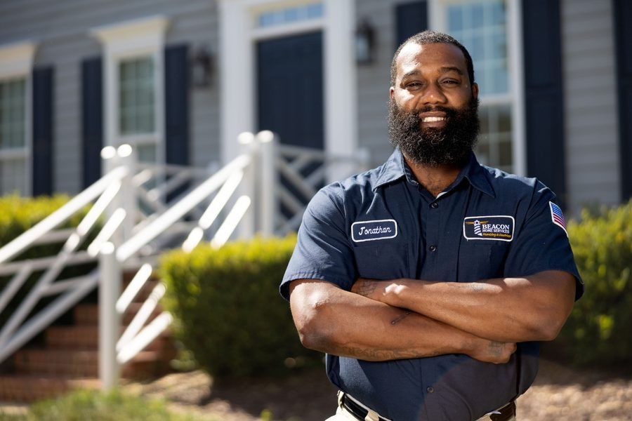 Bearded service technician with arms crossed, wearing a blue uniform with name patch 'Jonathan' outside a suburban home.