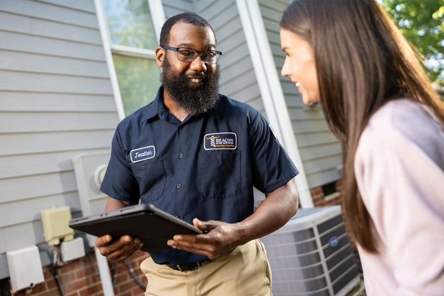 Bearded service technician named Jonathan in a navy shirt shows a tablet to a smiling woman outside a house.