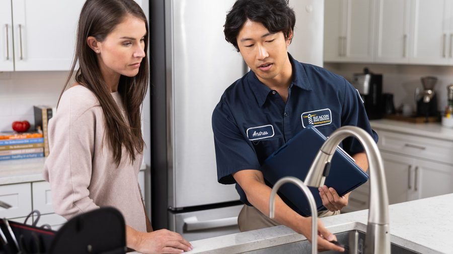 Technician named Andrew in a navy shirt shows a clipboard to a woman in a bright kitchen beside a fridge.